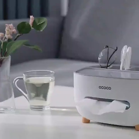 A 25-second product demo of the CORECOM® ECOCO Desk Organizer. The video shows a hand placing a smartphone, a TV remote, and a pair of glasses into the back slots, then pulling a single tissue from the front dispenser effortlessly. It highlights the sleek design from all angles and shows the product being placed back into its branded CORECOM® gift box.