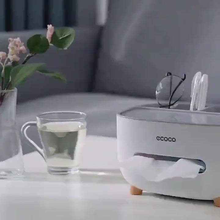 A 25-second product demo of the CORECOM® ECOCO Desk Organizer. The video shows a hand placing a smartphone, a TV remote, and a pair of glasses into the back slots, then pulling a single tissue from the front dispenser effortlessly. It highlights the sleek design from all angles and shows the product being placed back into its branded CORECOM® gift box.